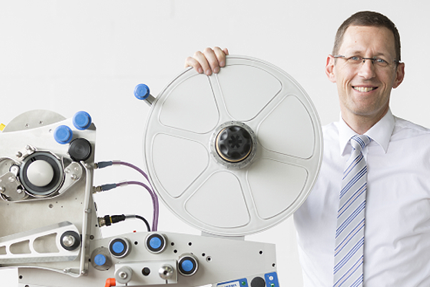 Ein Mann in einem Hemd und einer Krawatte hält eine große Scheibe und steht neben technischen Geräten mit Schläuchen und Anschlüssen.