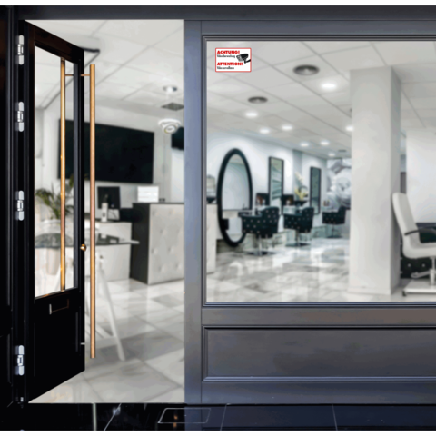 Glasfront eines Salons mit einer offenen Tür, die einen modernen Innenraum mit Friseurstühlen und Spiegeln zeigt. Ein Warnschild ist sichtbar.