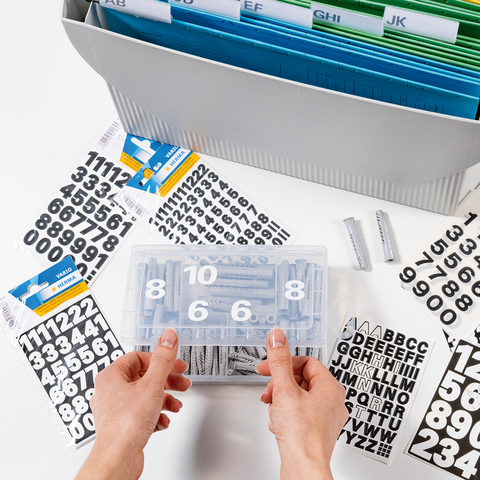 Hände halten eine durchsichtige Box mit Zahlen und Buchstaben. Im Hintergrund sind beschriftete Registerkarten und zusätzliche Zahlenblätter zu sehen.
