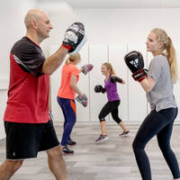 Eine Gruppe von Personen trainiert Boxen in einem Fitnessstudio. Ein Trainer hält Pratzen, während die Teilnehmer ihre Techniken üben.