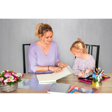 Eine Frau und ein Mädchen sitzen an einem Tisch und betrachten gemeinsam ein Buch. Auf dem Tisch liegen mehrere Schreibutensilien und ein Blumenstrauß.