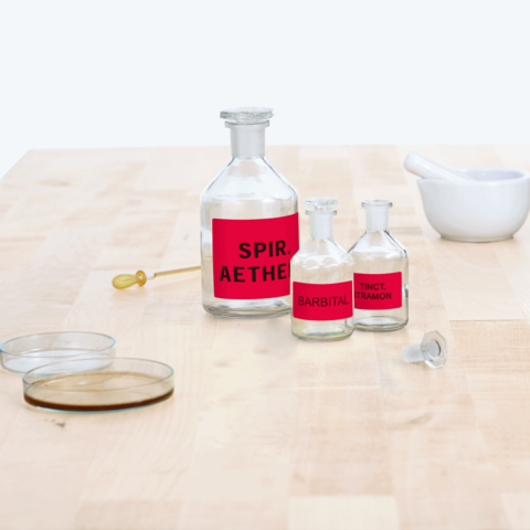 Flaschen mit roten Etiketten, beschriftet mit "Spir. Aether", "Barbital" und "Tinct. Tramon", auf einem Holztisch, daneben ein Mörser.