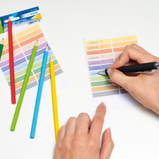 Bunte Stifte liegen neben einem Blatt, auf dem mit einem Stift geschrieben wird. Farbkarten sind ebenfalls sichtbar.