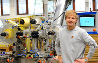 Ein Mann steht neben einer modernen Maschinenanlage zur Verarbeitung von Materialien, umgeben von technischen Geräten und Bildschirmen.