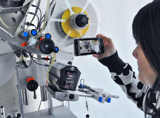 Eine Person hält ein Smartphone und fotografiert eine Maschine mit verschiedenen technischen Komponenten und Rollen.