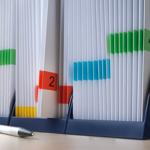 Ein Ordner mit bunten Markierungen und nummerierten Etiketten, der verschiedene Dokumente sortiert. Daneben liegt ein Stift.