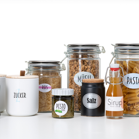 Gläser mit verschiedenen Lebensmitteln wie Reis, Müsli, Pasta, Pesto, Salz, Zucker und Sirup, alle mit beschrifteten Etiketten versehen.