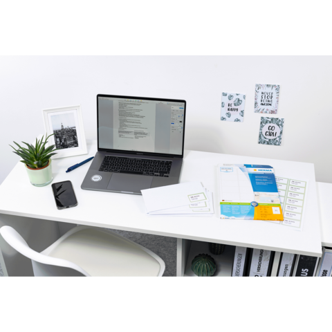 Schreibtisch mit Laptop, Smartphone und Pflanzen. Auf dem Tisch liegen Dokumente und Notizen, während an der Wand dekorative Bilder hängen.