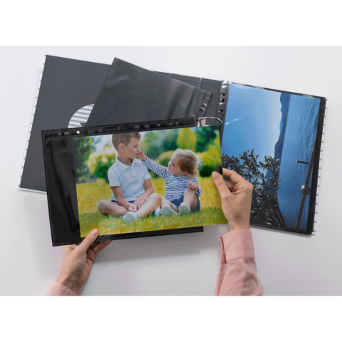 Eine Hand hält ein Foto von zwei Kindern, die im Freien auf dem Gras sitzen. Im Hintergrund sind weitere Fotos in einem Album sichtbar.