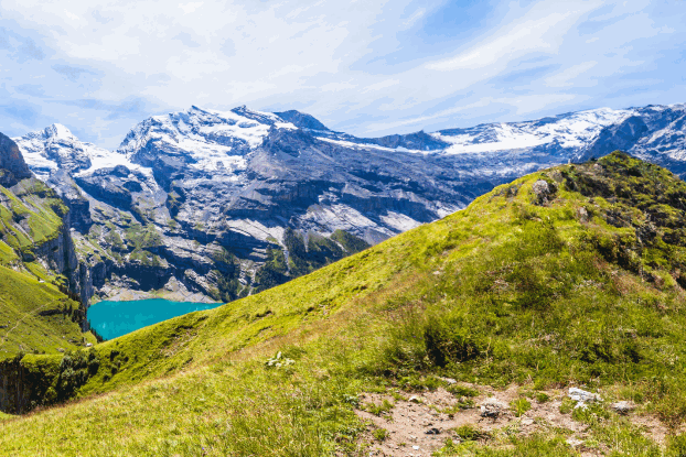 Panoramablick auf saftig grüne Hügel und schneebedeckte Berge, umgeben von klarem Wasser und strahlend blauem Himmel.