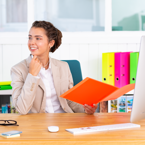 Eine Frau in einem Büro sitzt am Schreibtisch, hält einen orangefarbenen Ordner und lächelt, während sie nachdenklich aussieht.