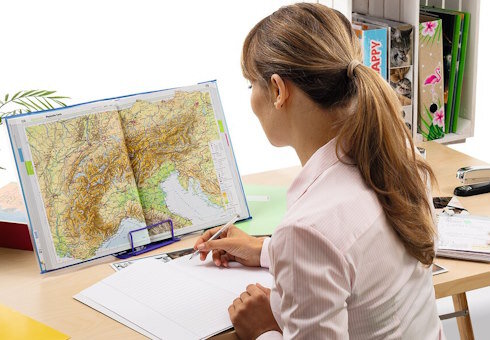 Frau steht vor einem geöffneten Atlas auf einem Schreibtisch.