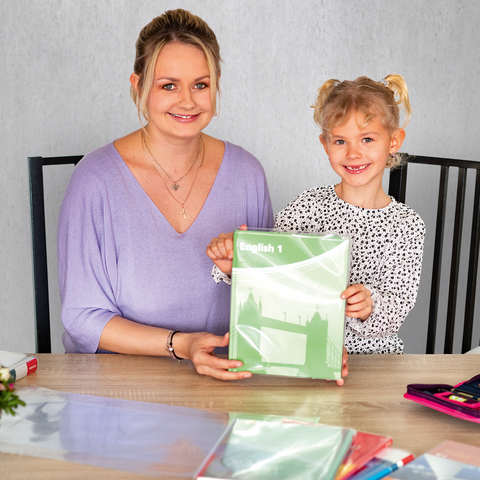 Eine Frau und ein Mädchen sitzen an einem Tisch, halten ein grünes Heft und lächeln. Auf dem Tisch liegen Schreibmaterialien und Blumen.