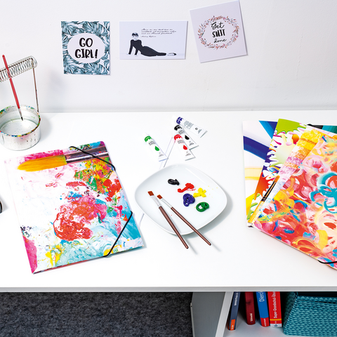 Ein Tisch mit Malutensilien, darunter Aquarellfarben, Pinsel, Papier und ein Holzmodell. An der Wand hängen inspirierende Zitate.