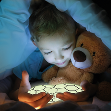Ein Kind sitzt im Bett, hält ein leuchtendes Objekt in den Händen und umarmt einen Teddy, während es unter einer Decke liegt.