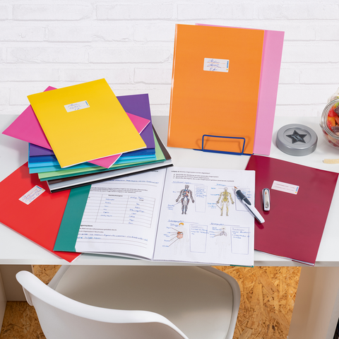 Bunte Ordner und Unterlagen auf einem Schreibtisch, daneben ein Glas mit Süßigkeiten und Bürobedarf wie einen Hefter und einen Stift.