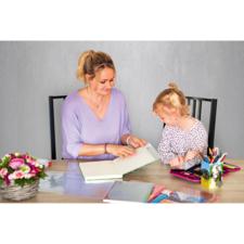 Eine Frau und ein Mädchen sitzen an einem Tisch und betrachten gemeinsam ein Buch. Auf dem Tisch liegen verschiedene Schreibwaren und Blumen.