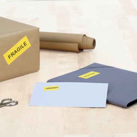 Ein Karton mit dem Aufkleber "FRAGILE", daneben ein graues Kuvert mit dem Aufkleber "DRINGEND!" und eine Schere auf einem Holztisch.