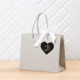 Graue Einkaufstasche mit einem schwarzen Herzaufkleber, der die Zahl 25 zeigt, und einer weißen Schleife.