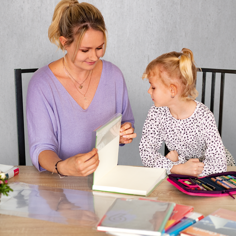 Eine Frau und ein kleines Mädchen sitzen an einem Tisch, während sie gemeinsam an einem Kunstprojekt arbeiten. Bunte Stifte und Materialien sind sichtbar.
