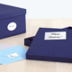 Eine geöffnete, dunkelblaue Geschenkbox mit einem "Happy Birthday!"-Aufkleber und zwei Umschlägen in hellblau und grau daneben.