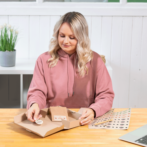 Eine Person mit lockigem Haar in einem rosa Hoodie öffnet eine Pappverpackung und betrachtet einen Aufkleber auf einem Tisch.