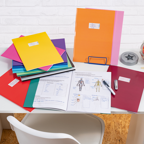 Bunte Ordner und Dokumente auf einem Schreibtisch, neben einem Glas mit Süßigkeiten und Bürobedarf wie einem Hefter und einem Stift.