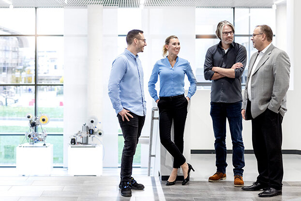 Vier Personen in einem modernen Büro unterhalten sich, während im Hintergrund ein Industriegebäude und eine weitläufige Anlage zu sehen sind.