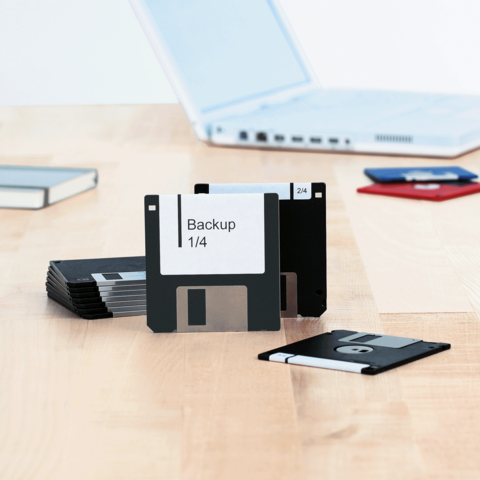 Mehrere Disketten liegen auf einem Tisch, eine zeigt das Etikett "Backup 1/4". Im Hintergrund ist ein Laptop sichtbar.