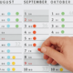 Ein Hand, die einen roten Punkt auf einem Kalenderblatt für September und Oktober anzeigt, mit markierten Tagen und Wochentagen.