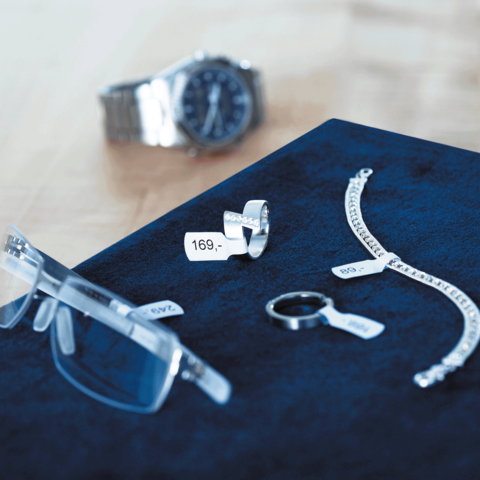 Eine Uhr, eine Brille, ein Ring, ein Armband und ein Schlüsselring liegen auf einem blauen Untergrund, jeweils mit Preisschildern versehen.