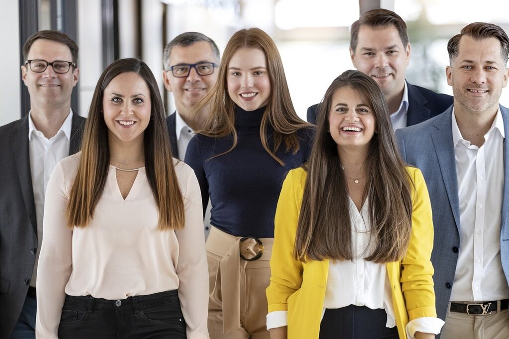 Eine Gruppe von sieben Personen steht in einem Flur, lächelnd und in formeller Kleidung, einige tragen Hemden und Anzüge.