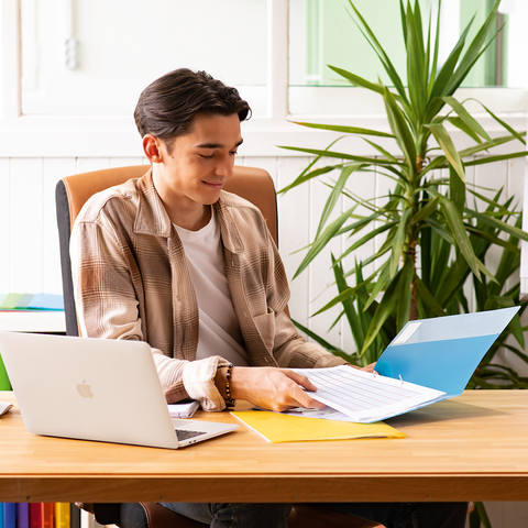 Junger Mann sitzt an einem Schreibtisch, arbeitet mit einem Laptop und blättert durch Dokumente in einer bunten Büroumgebung.