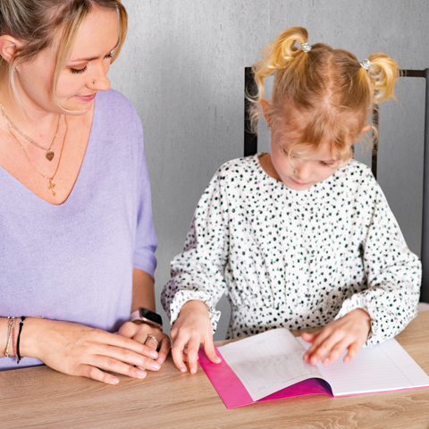 Eine Frau sitzt an einem Tisch neben einem Mädchen, das in einem Notizbuch schreibt. Beide konzentrieren sich auf die Aktivität.