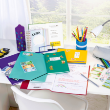 Ein Schreibtisch mit bunten Ordnern, Schreibmaterialien, einem Laptop und einer Lampe. Ein Fenster im Hintergrund lässt Tageslicht herein.