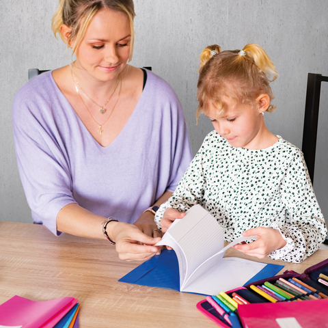 Ein Kind und ein Jugendlicher sitzen an einem Schreibtisch und zeichnen. Im Hintergrund steht ein Regal mit Spielzeug und Büchern.