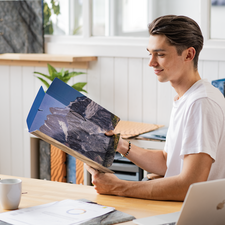 Ein junger Mann sitzt an einem Tisch und betrachtet ein aufgeschlagenes Buch mit Landschaftsbildern. Eine Tasse steht daneben.