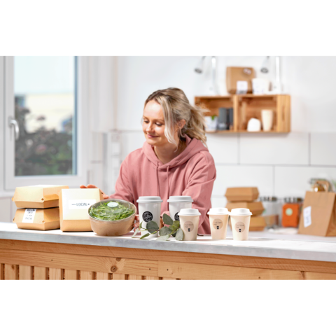 Eine Frau in einem rosa Hoodie steht an einem Tisch mit Essensverpackungen, einer Schüssel Salat und Kaffeebechern in einer modernen Küche.