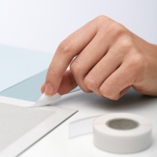 Eine Hand zieht ein Stück Klebeband von einer grauen Oberfläche ab, während ein weiteres Stück Klebeband daneben liegt.