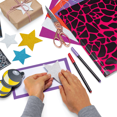 Hände, die einen funkelnden Stern auf ein buntes Papier kleben, umgeben von Bastelmaterialien wie Scheren, Stiften und Geschenkpapier.