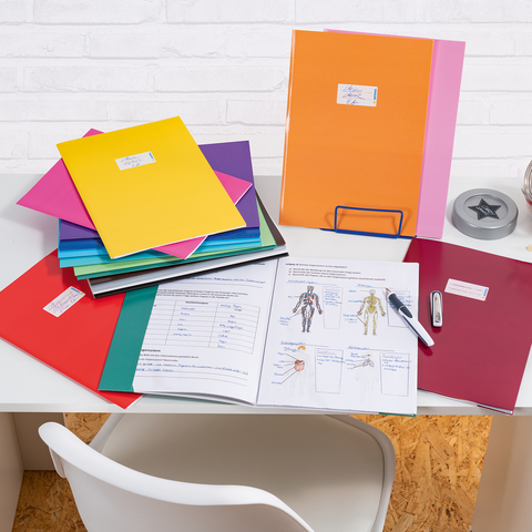 Bunte Ordner und Unterlagen auf einem Schreibtisch, daneben ein Glas mit Süßigkeiten und Bürobedarf wie einen Hefter und einen Stift.