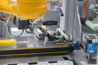 Industrieller Roboterarm mit Greifmechanismus, der Objekte auf einem Förderband verarbeitet. Technische Komponenten und Sensoren sind gut sichtbar.