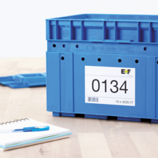 Blaue Kunststoffbox mit der Aufschrift "0134" und einem Etikett, daneben ein Notizblock und ein Stift auf einem Tisch.