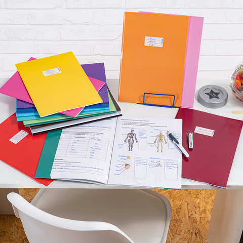 Bunte Ordner und Dokumente auf einem Schreibtisch, daneben ein Glas mit Süßigkeiten und Bürobedarf wie ein Hefter und ein Stift.