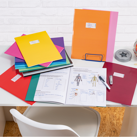 Bunte Ordner und Dokumente auf einem Schreibtisch, daneben ein Glas mit Süßigkeiten und Bürobedarf wie einen Hefter und einen Stift.