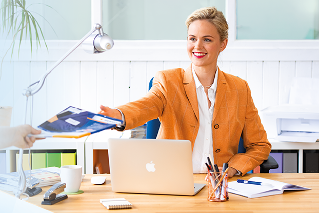 Zwei Personen sitzen an einem Tisch und präsentieren Etiketten. Links ist ein Mann mit Etikettenblättern, rechts eine Frau mit einem Laptop und Etiketten.