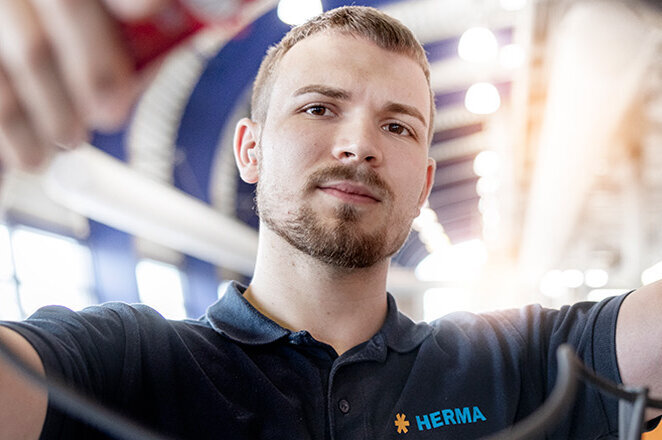 Ein junger Mann in Arbeitskleidung hält Kabel in einer modernen Umgebung mit einem Gebäude und Bäumen im Hintergrund.
