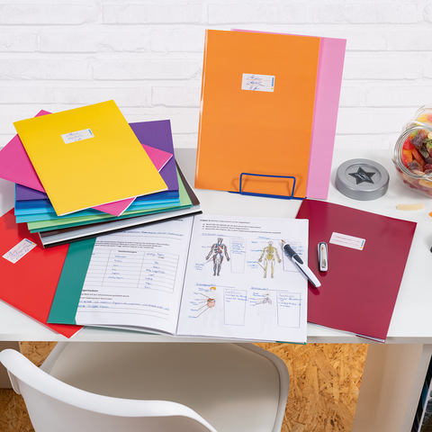 Bunte Ordner und Unterlagen auf einem Schreibtisch, daneben ein Glas mit Süßigkeiten und Bürobedarf wie ein Hefter und ein Stift.