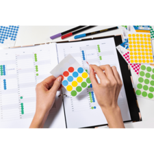 Eine Person hält ein Blatt mit bunten Aufklebern über einem geöffneten Planer, während sie die Seiten mit Markierungen betrachtet.