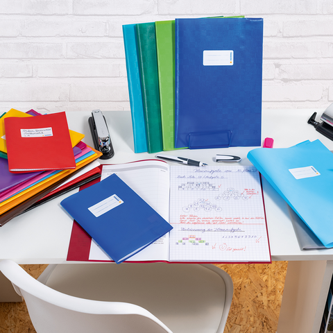 Bunte Ordner und Notizhefte auf einem Schreibtisch, dazu ein Hefter, ein Stift und ein offenes Notizbuch mit handschriftlichen Notizen.